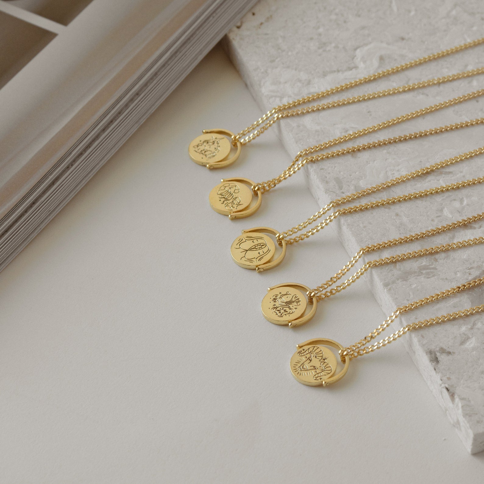 Five Zodiac Spinner Necklaces with astrological designs and personalized engraving are displayed on a white stone surface next to a book.