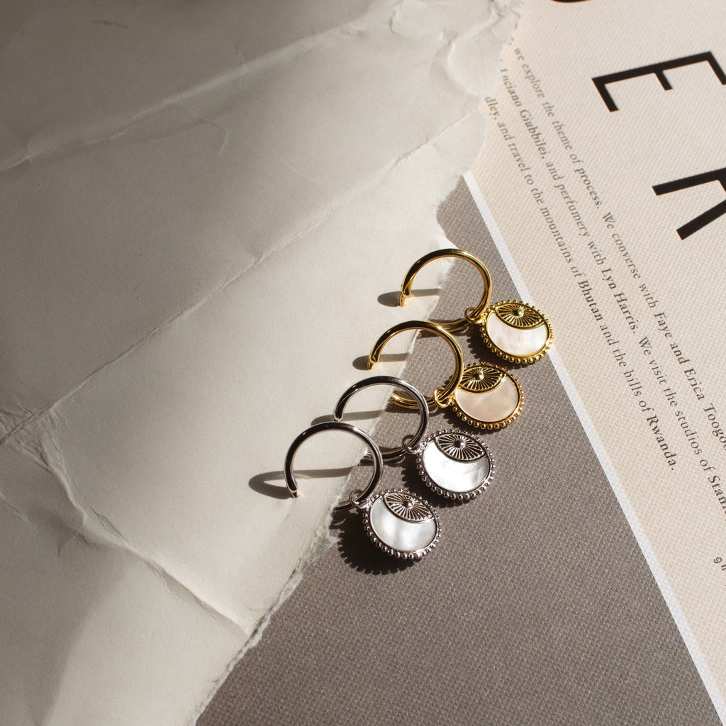 Two pairs of Pia Pearl Coin Hoops, featuring gold and silver sunburst designs, are displayed on a textured neutral background with paper.