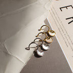 Two pairs of Pia Pearl Coin Hoops, featuring gold and silver sunburst designs, are displayed on a textured neutral background with paper.