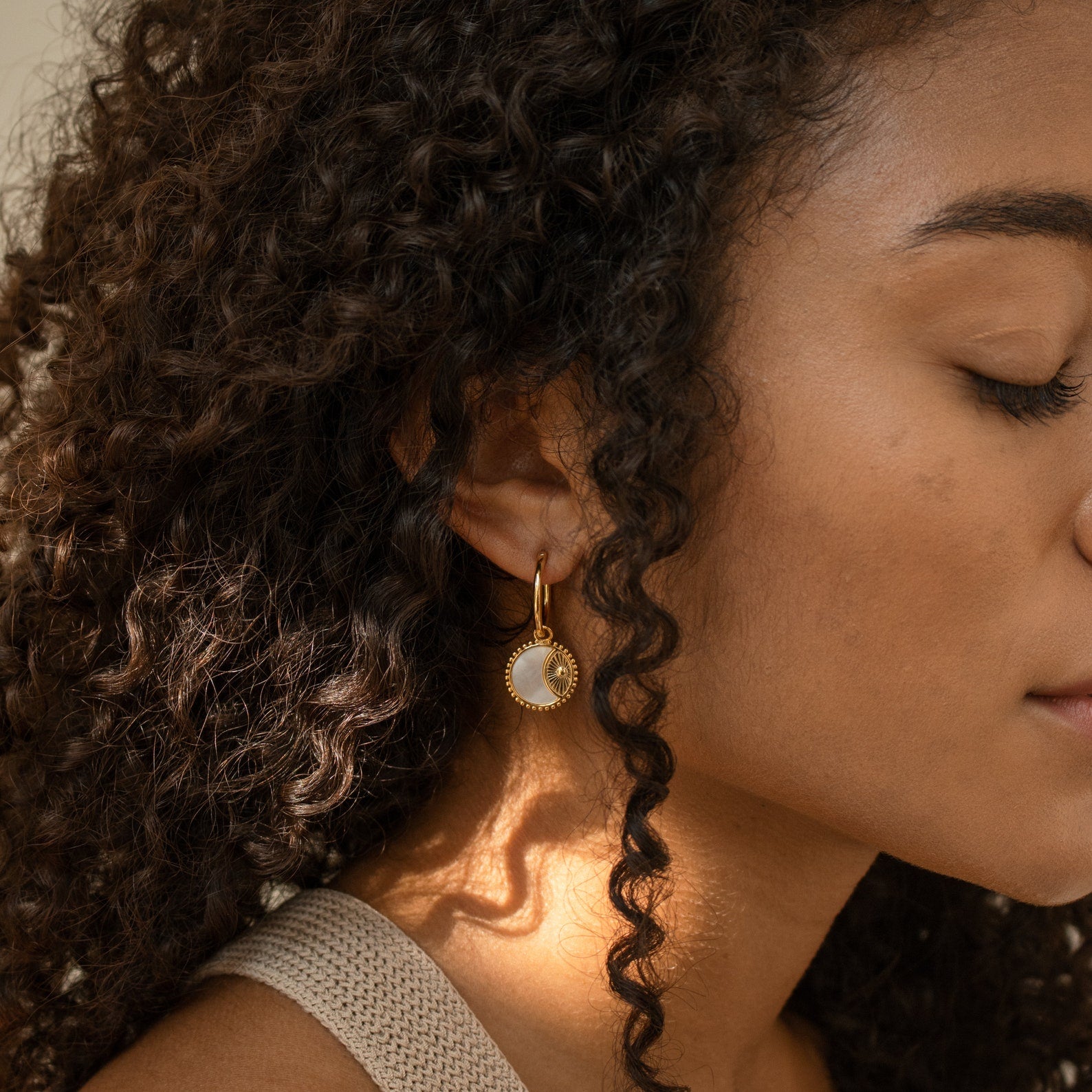Woman with curly hair wears Pia Pearl Coin Hoops in gold and white, eyes closed, bathed in soft natural light.