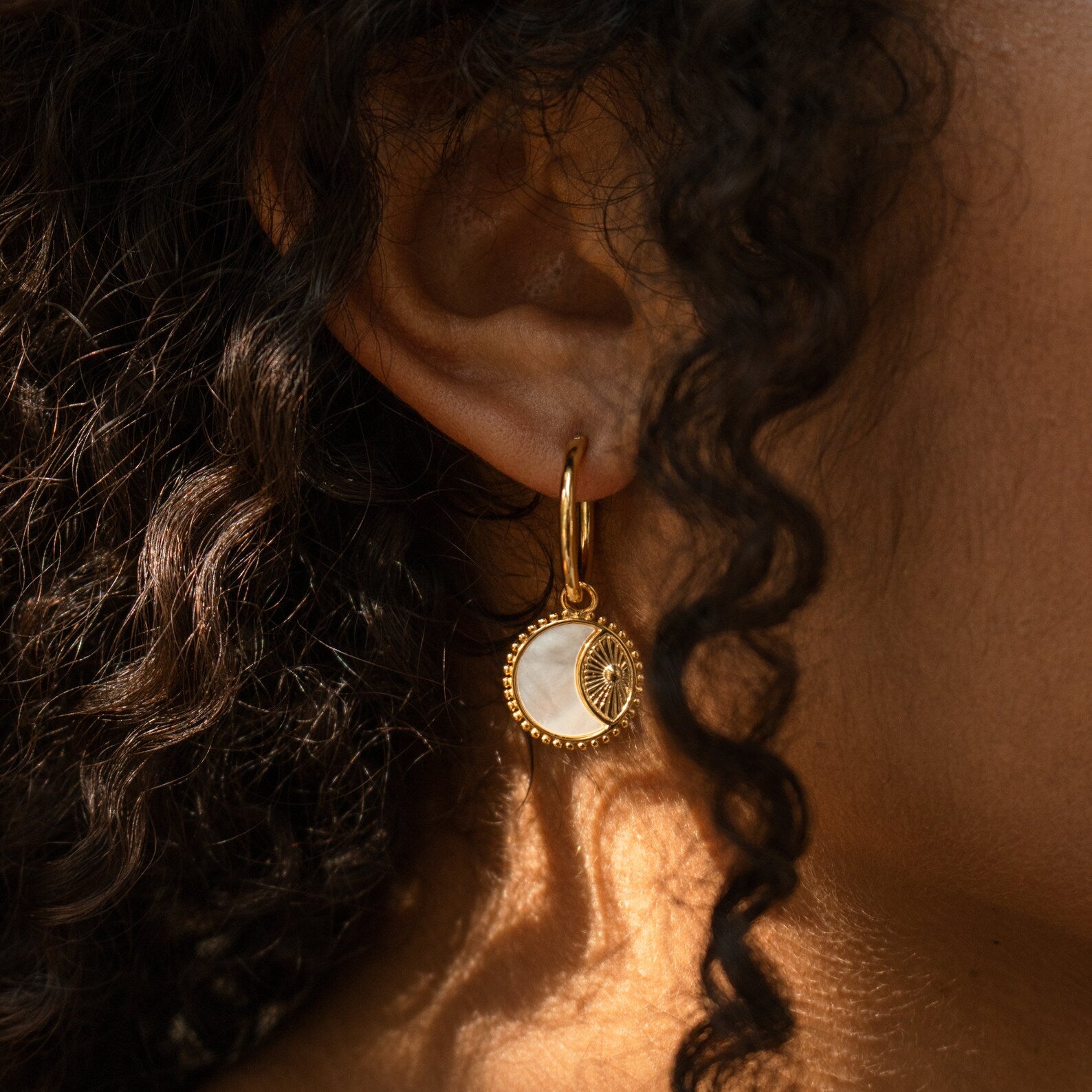 Close-up of an ear wearing Pia Pearl Coin Hoops—gold hoop earrings with a round, white and gold sunburst coin pendant—ideal for boho earring lovers.