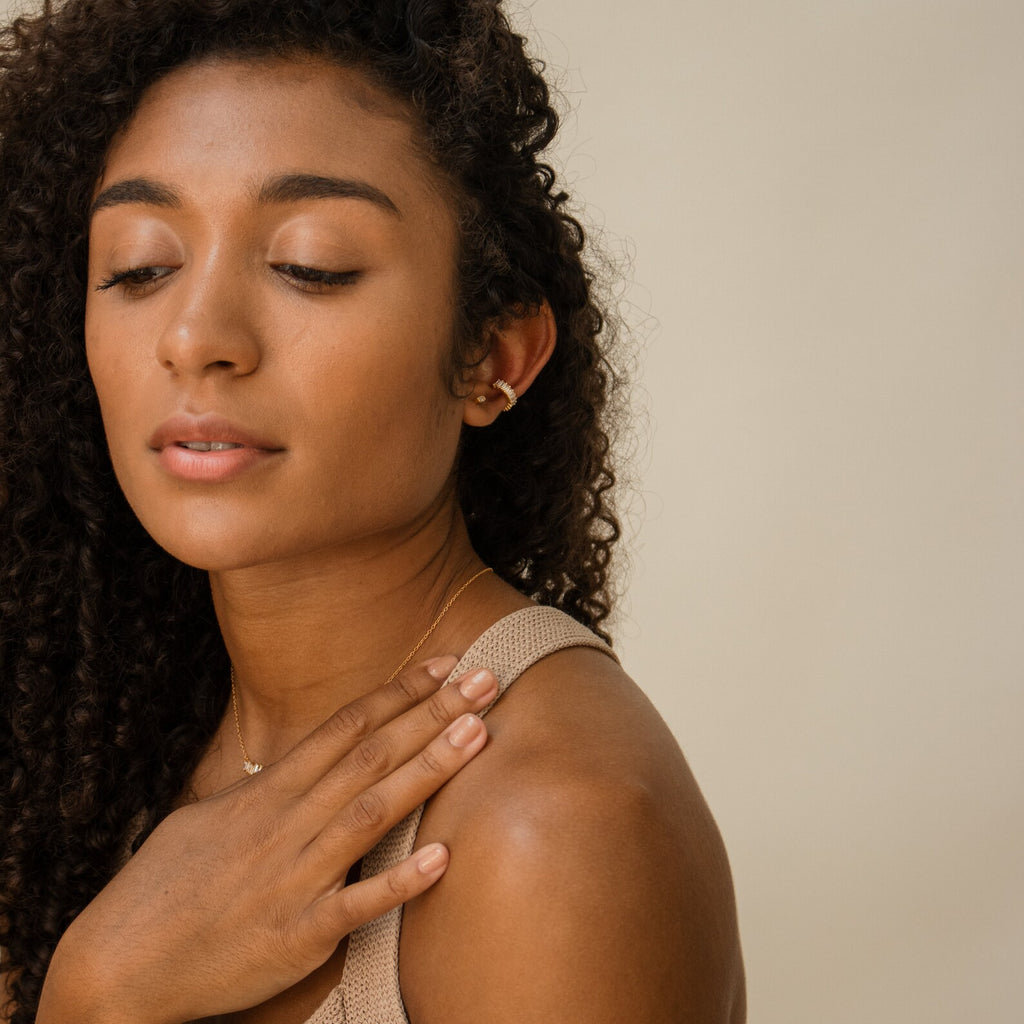 A woman with curly hair in a beige top looks down calmly, touching her shoulder and showcasing the delicate Baguette Ear Cuffs.