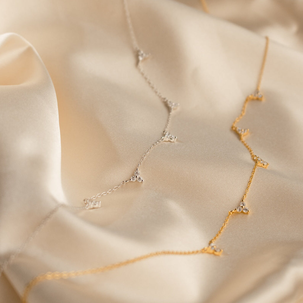 The Hana Diamond Station Necklace, a delicate gold and silver constellation design, is elegantly displayed on smooth, light beige satin fabric.