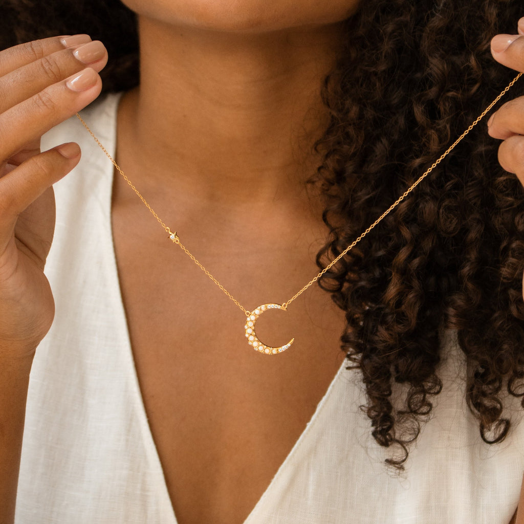 A woman with curly hair, wearing a white top, showcases the Opal Crescent Moon Necklace in gold—an ideal boho accessory.