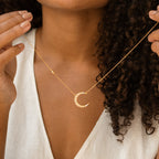 A woman with curly hair, wearing a white top, showcases the Opal Crescent Moon Necklace in gold—an ideal boho accessory.