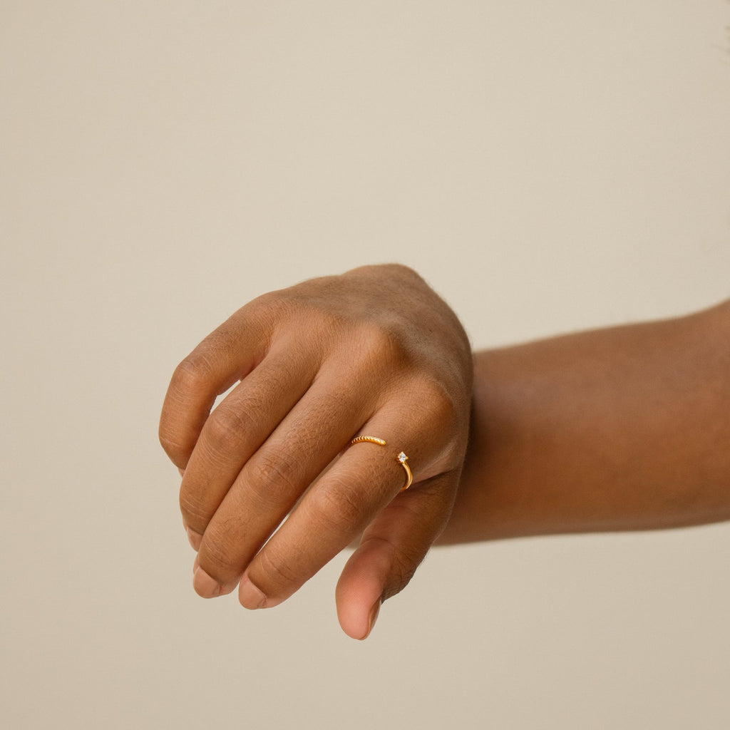 A hand showcases the Cecilia Pave Ring, featuring a small stone, set against a plain beige background.