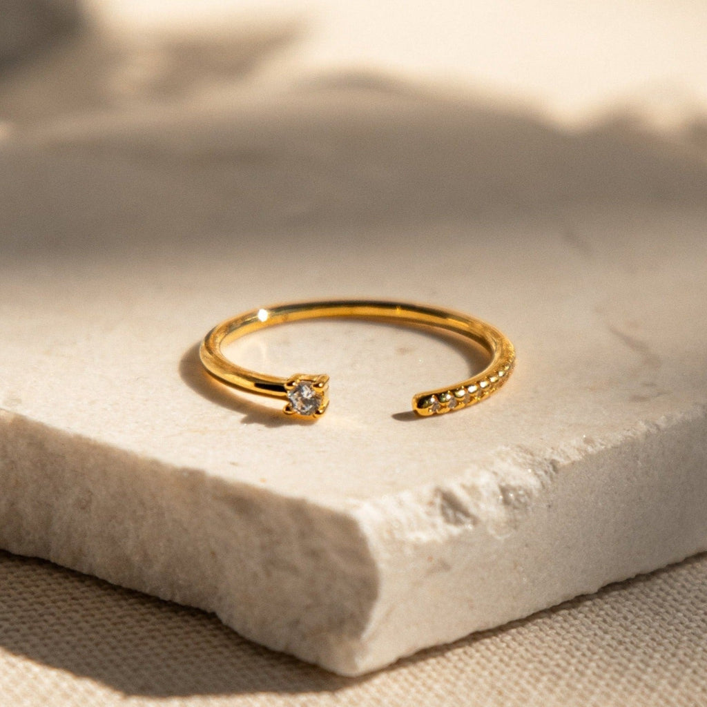 The Cecilia Pave Ring is a gold open-style ring with a small central diamond and a delicate pavé row of tiny stones, displayed on a textured stone surface.