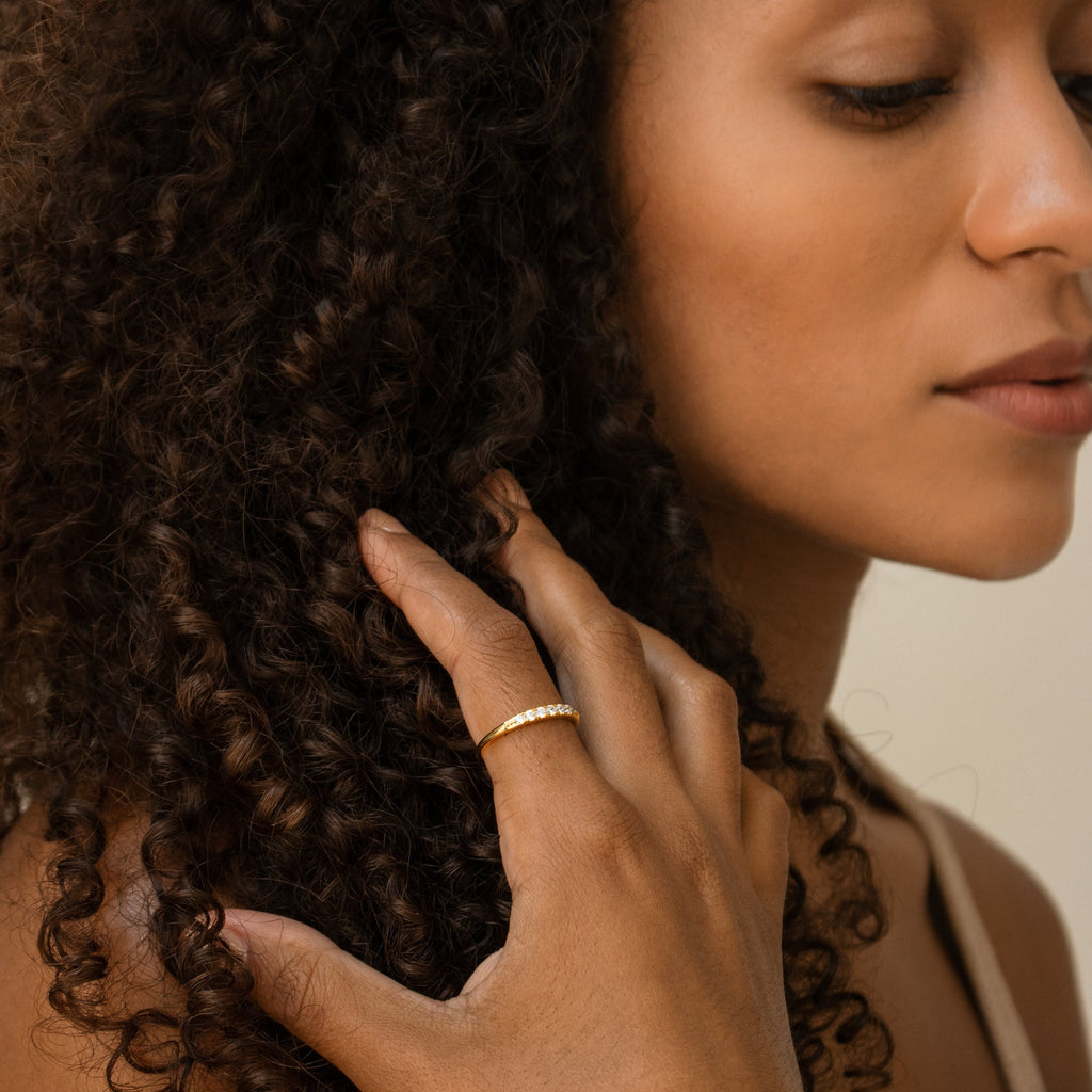 A woman with curly hair touches her hair, wearing a simple gold ring and the Lace Pave Ring—a delicate, thin diamond band—on her finger.