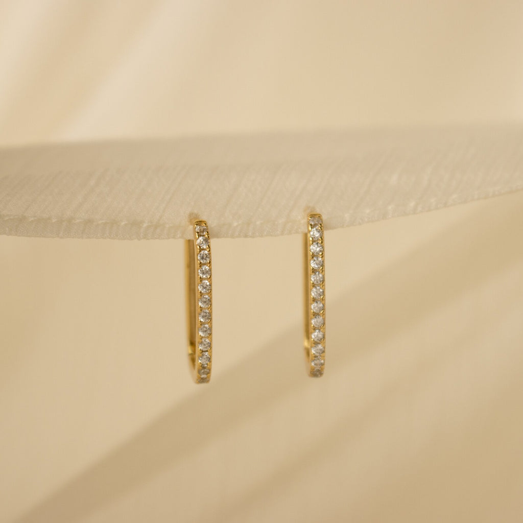 Displayed on a textured light beige surface, the Pave Link Earrings feature minimal gold hoops adorned with sparkling diamonds.