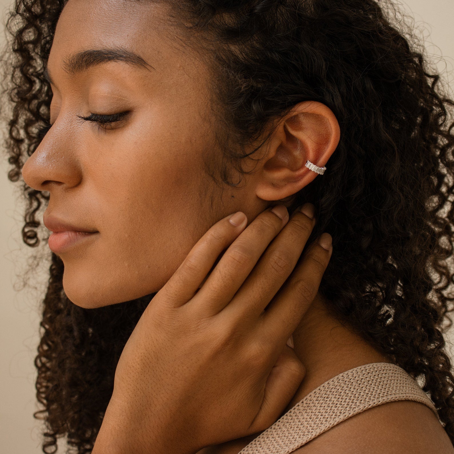 A woman with curly hair, eyes closed, gently touches her neck and ear while wearing Baguette Ear Cuffs in silver.