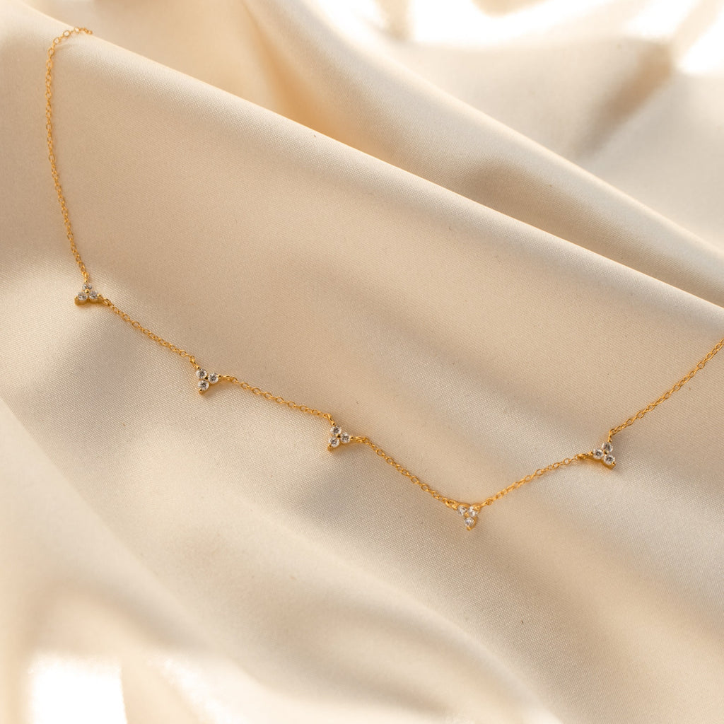 The Hana Diamond Station Necklace features five delicate diamond charms on gold, perfect for layering. Shown on soft, light beige satin.