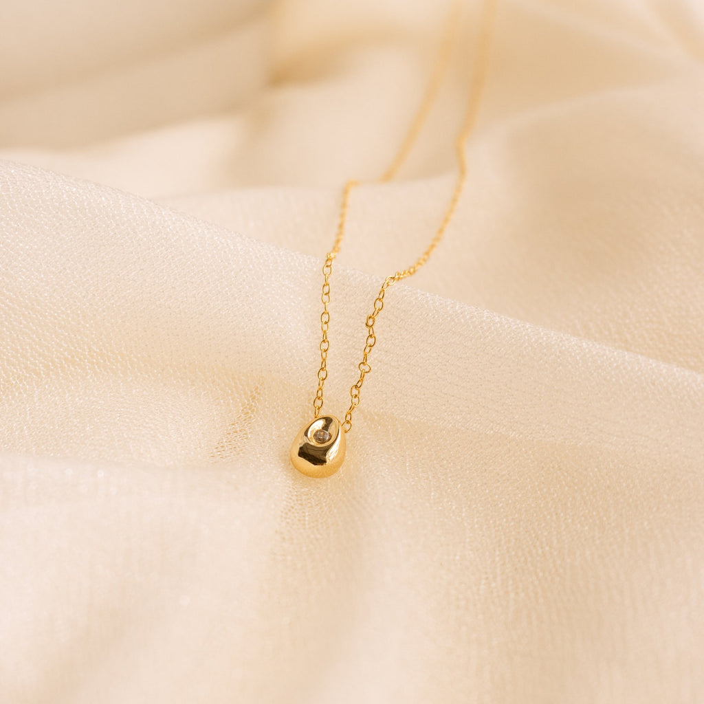 The Nugget Necklace features a delicate gold nugget pendant with a small gemstone, beautifully showcased on soft, beige fabric.