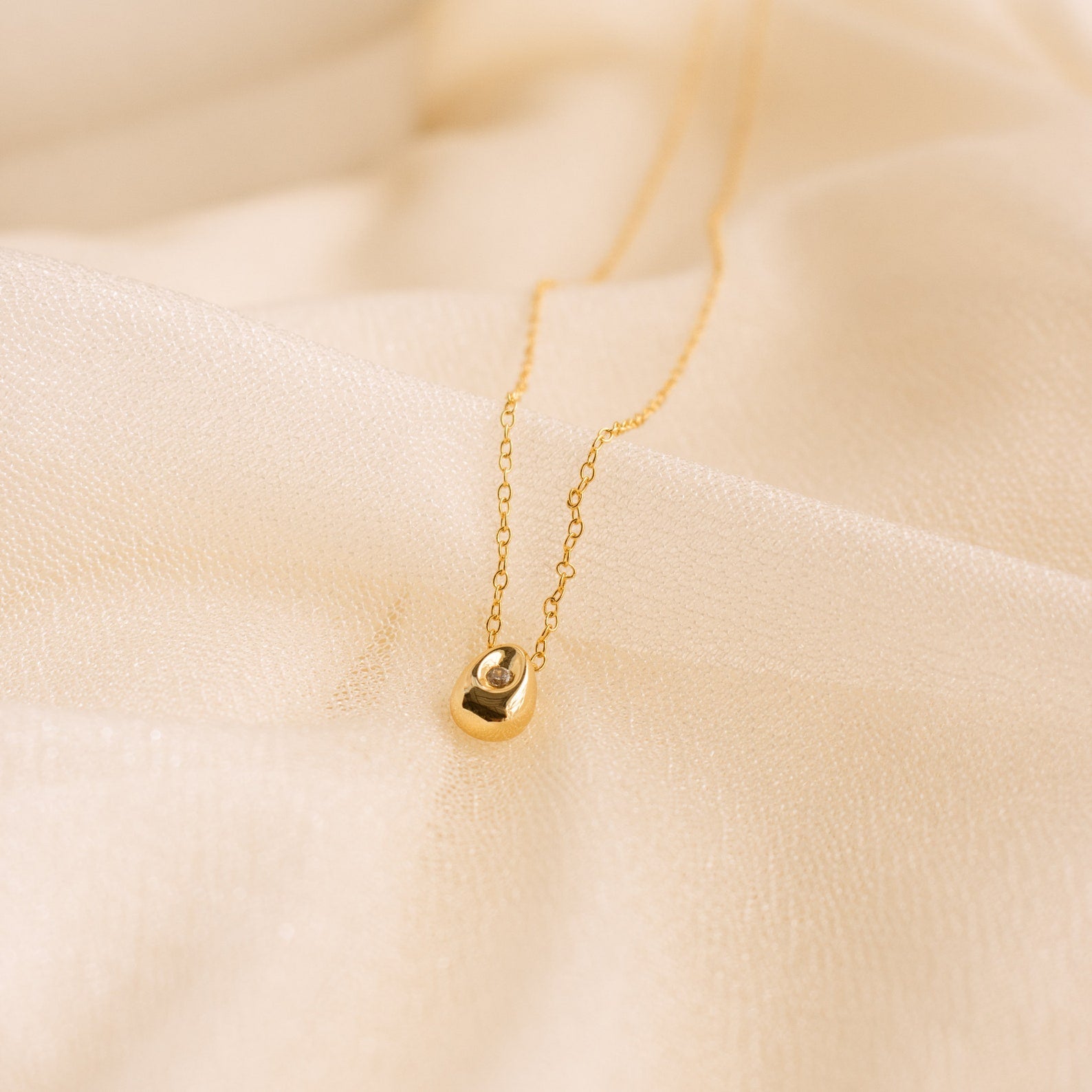 The Nugget Necklace features a delicate gold nugget pendant with a small gemstone, beautifully showcased on soft, beige fabric.