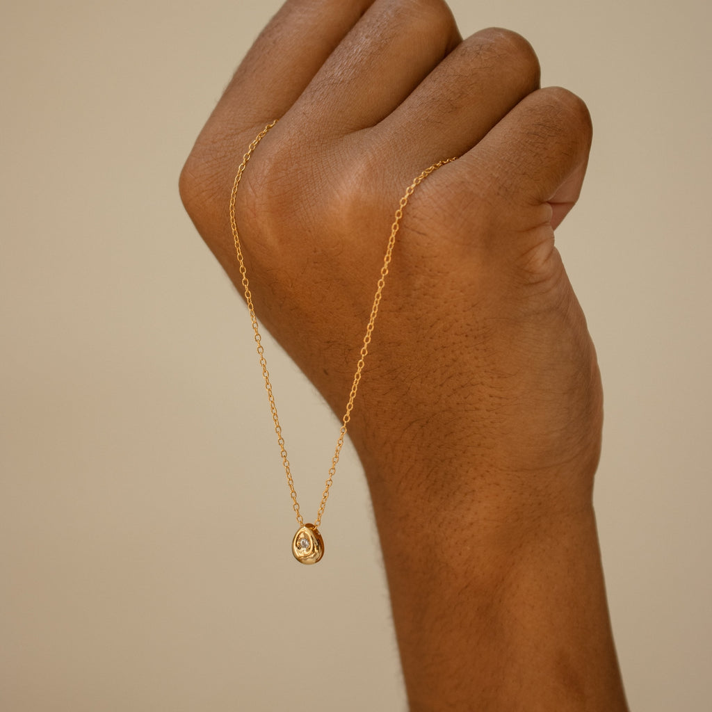 A hand holds up the Nugget Necklace, featuring a dainty round pendant, against a neutral background.