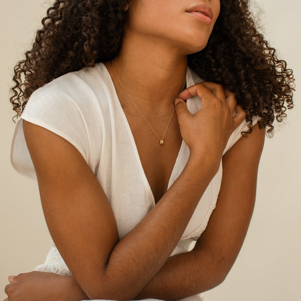 A woman in a white dress sits with her hand on her shoulder, wearing the Nugget Necklace.