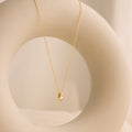 Minimal gold necklace with a teardrop-shaped pendant featuring a single inset stone, draped over a smooth circular ceramic sculpture in a softly lit neutral setting.