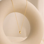 Minimal gold necklace with a teardrop-shaped pendant featuring a single inset stone, draped over a smooth circular ceramic sculpture in a softly lit neutral setting.