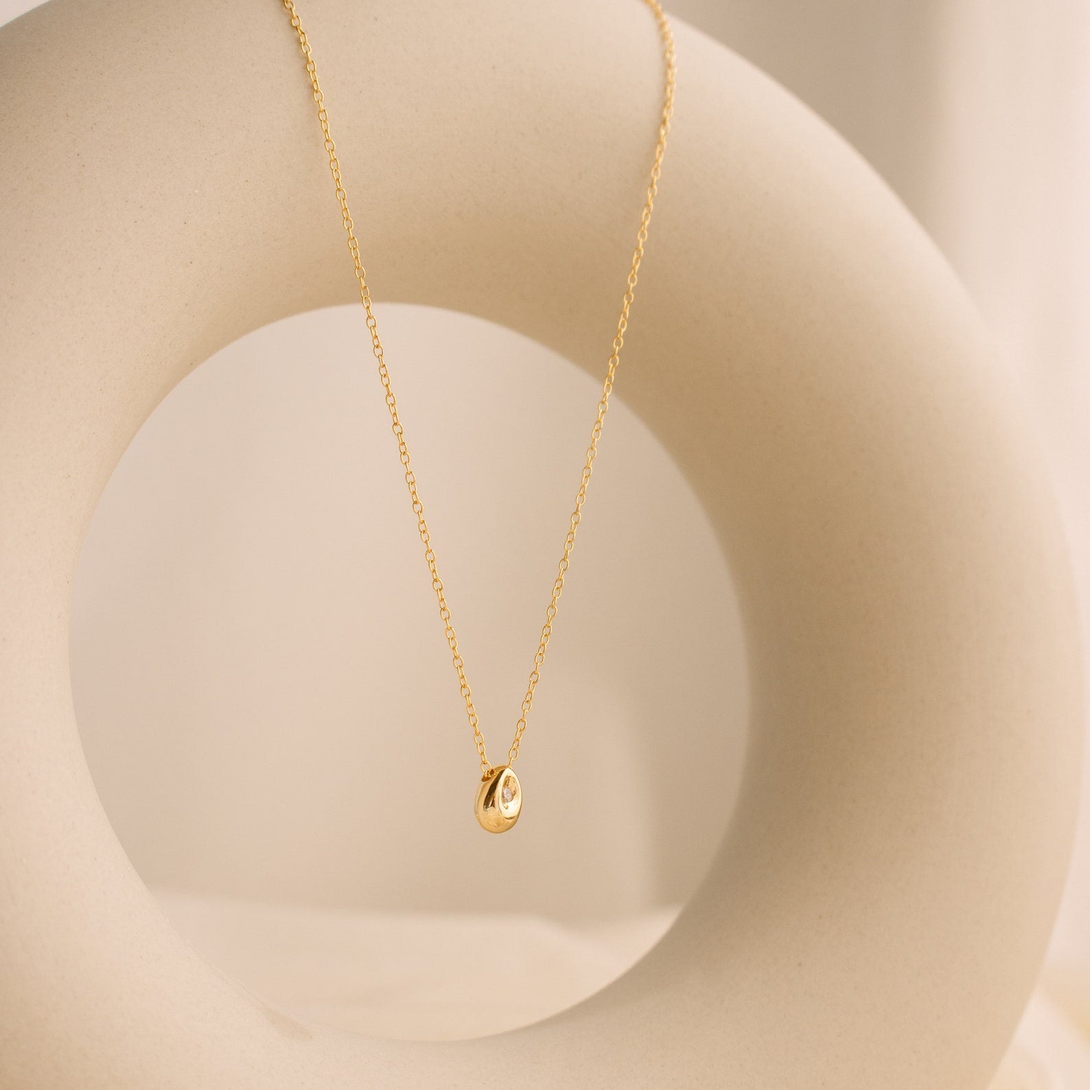 Minimal gold necklace with a teardrop-shaped pendant featuring a single inset stone, draped over a smooth circular ceramic sculpture in a softly lit neutral setting.
