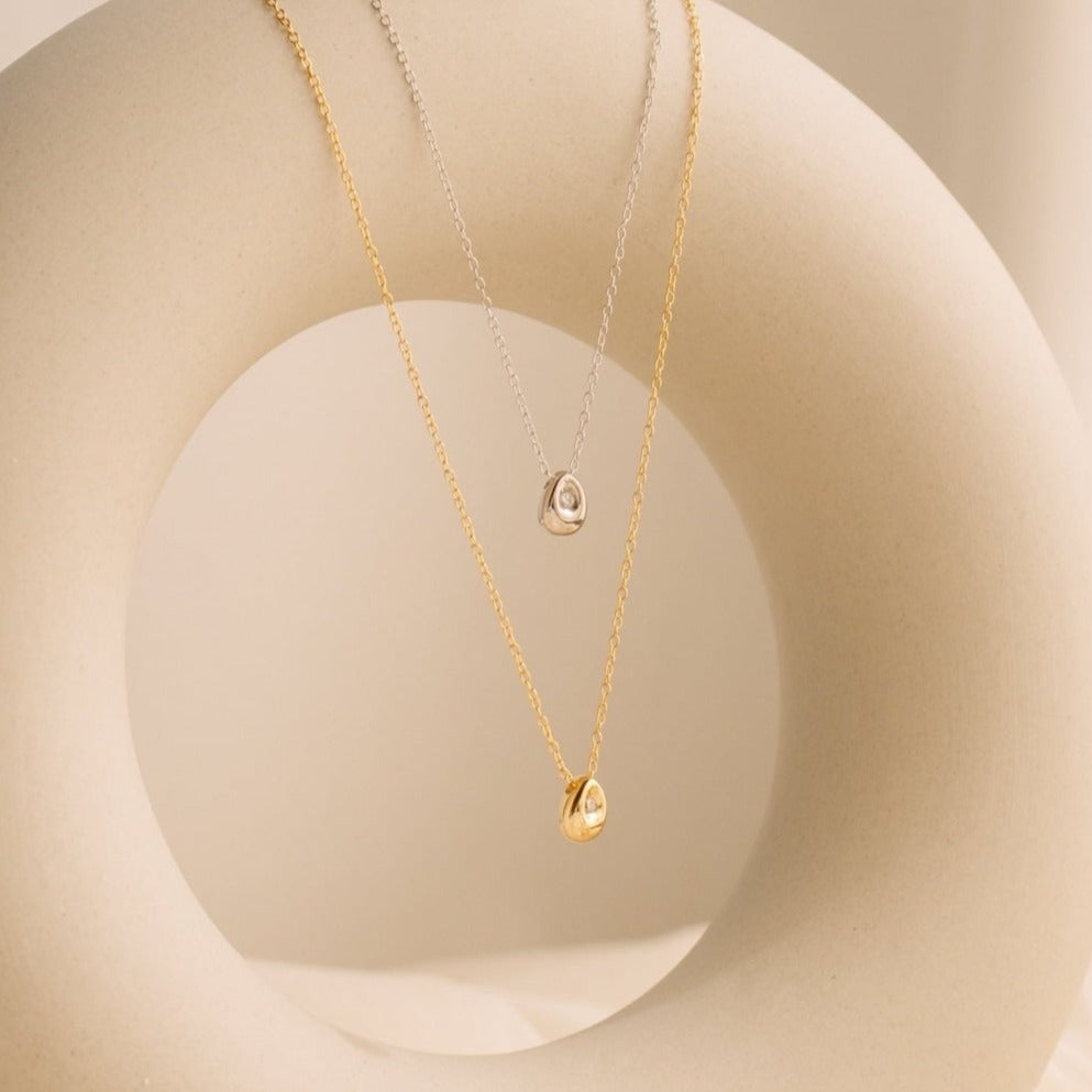 Two Nugget Necklaces—one gold, one silver—each with a delicate pendant, hang elegantly on a smooth circular display, making them the perfect pair for lovers of dainty necklaces.