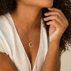 A woman, dressed in a white blouse, showcases the Opal Crescent Moon Necklace as she rests her hand near her chin.