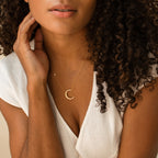 A woman with curly hair in a white blouse touches her neck while wearing the Opal Crescent Moon Necklace—a delicate boho piece featuring a gold crescent moon pendant.