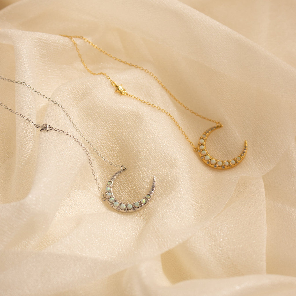 Two Opal Crescent Moon Necklaces—one silver, one gold—rest on soft cream fabric. This boho-inspired duo adds celestial charm to any look.