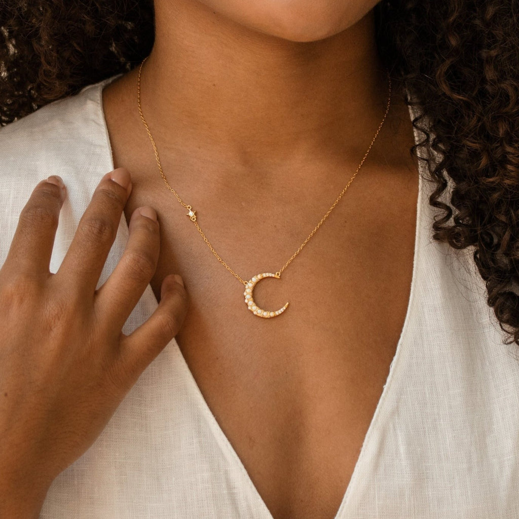 Gold crescent moon necklace adorned with pearl-like stones, worn by a model in a white V-neck top, with the charm resting just below the collarbone.