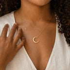 Gold crescent moon necklace adorned with pearl-like stones, worn by a model in a white V-neck top, with the charm resting just below the collarbone.