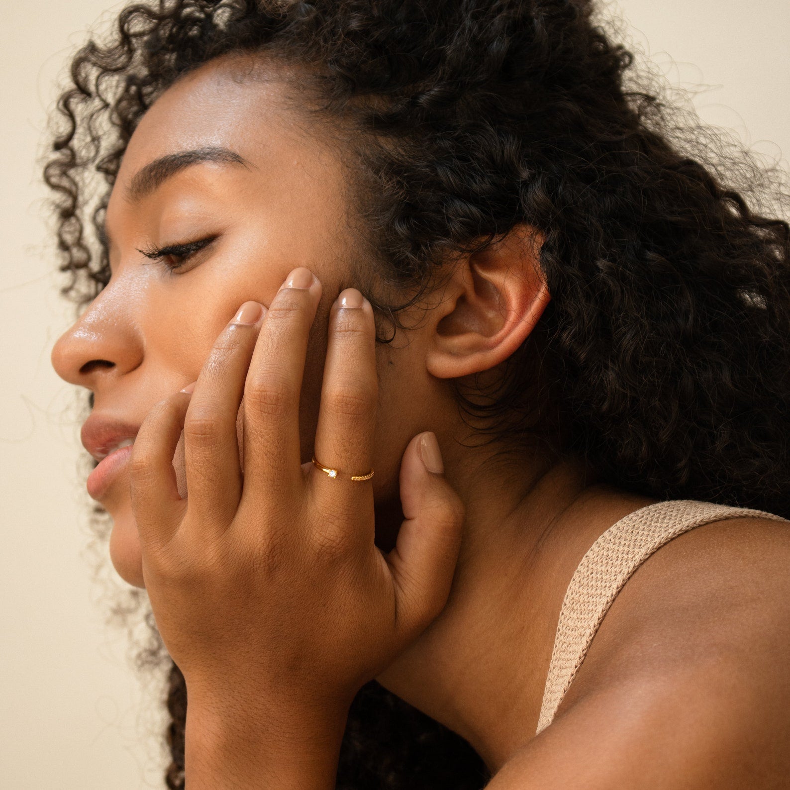 A woman with curly hair, dressed in neutral tones, rests her hand on her face, showcasing the Cecilia Pave Ring.