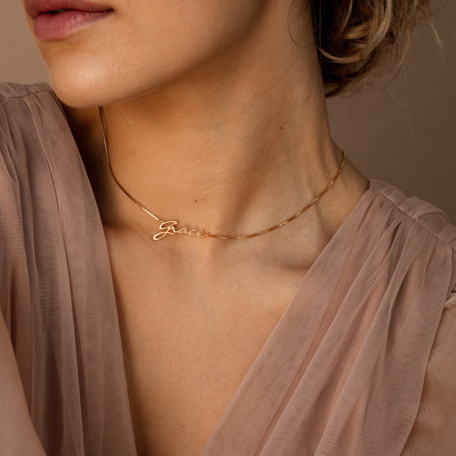A woman wears the Callie Box Chain Name Necklace in 18K Gold personalized with "Grace," paired with a sheer blush pink top.