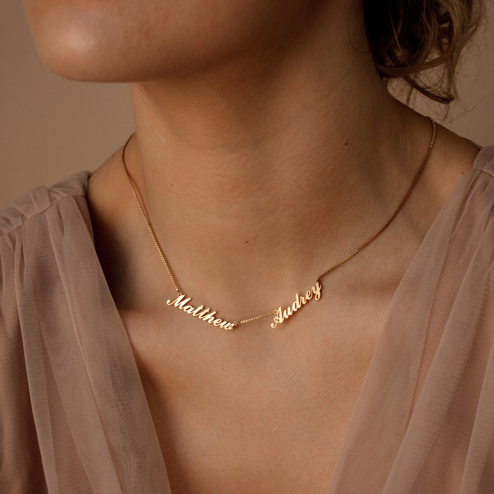 The Carrie Multiple Name Necklace in Box Chain, featuring the names Matthew and Audrey, rests on a woman's neck, perfectly complementing her light pink blouse. Experience the charm of personalized jewelry with this elegant piece.