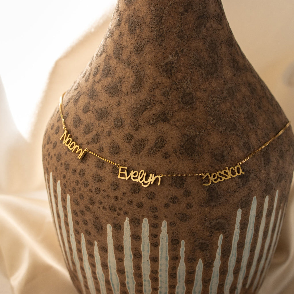 The Preppy Multiple Name Necklace in Box Chain, featuring Naomi, Evelyn, and Jessica in a youth-inspired font, is draped over a textured brown vase.