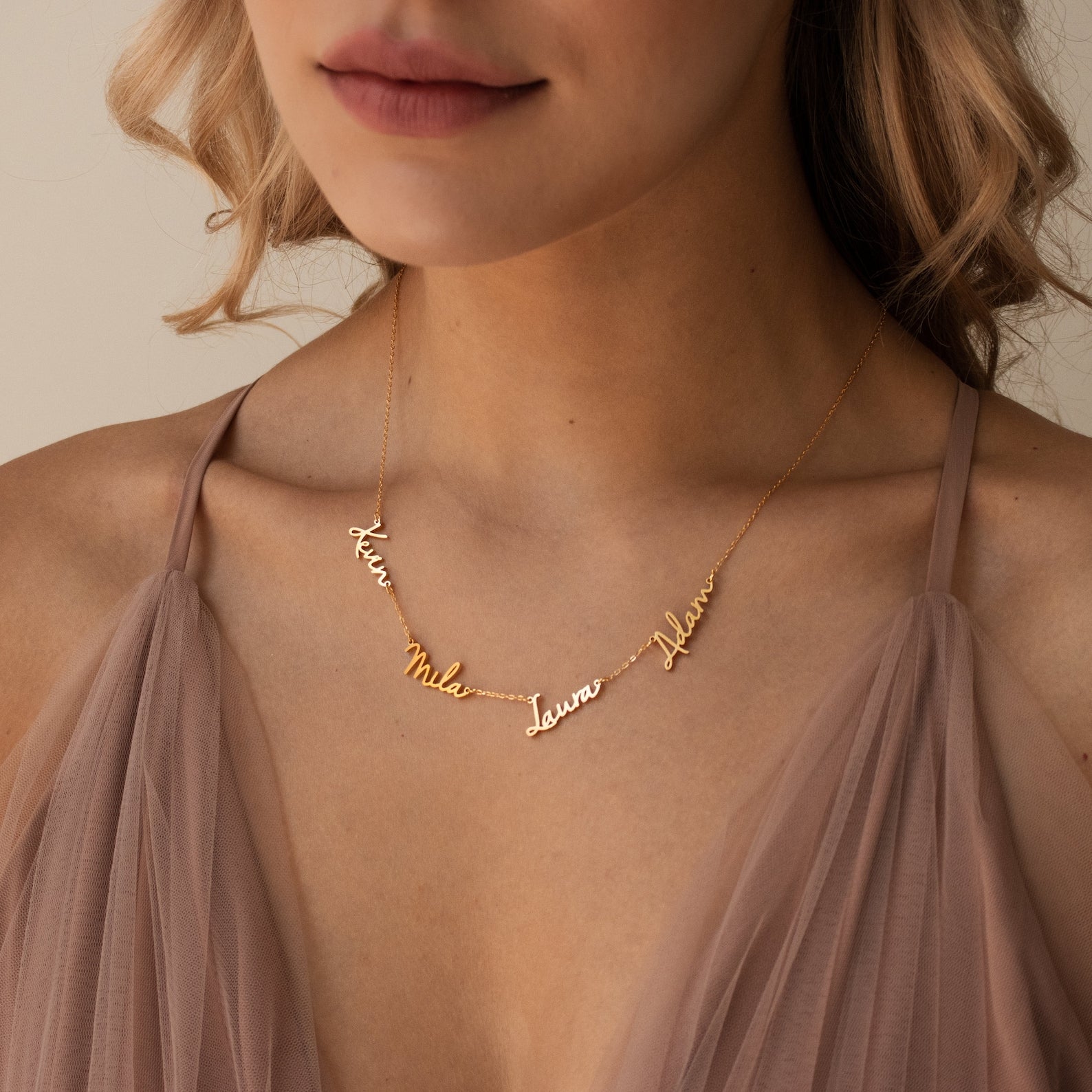 A woman wears the Nella Multiple Name Necklace in gold, customized with the names Kim, Mila, Laura, and Adam in elegant cursive, paired with a taupe dress.