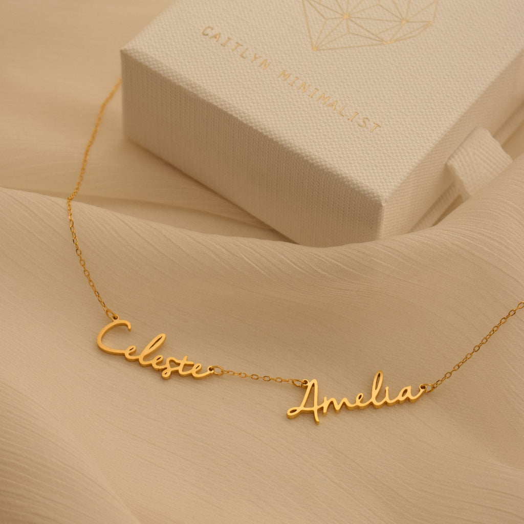 Nella Multiple Name Necklace in gold, featuring Celeste and Amelia script pendants, shown beside a textured white jewelry box.