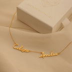 Nella Multiple Name Necklace in gold, featuring Celeste and Amelia script pendants, shown beside a textured white jewelry box.