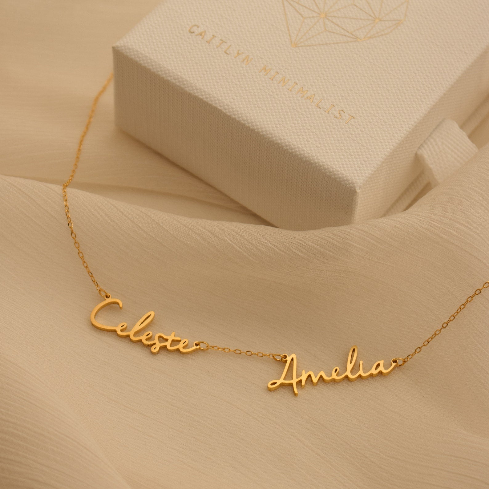 Nella Multiple Name Necklace in gold, featuring Celeste and Amelia script pendants, shown beside a textured white jewelry box.