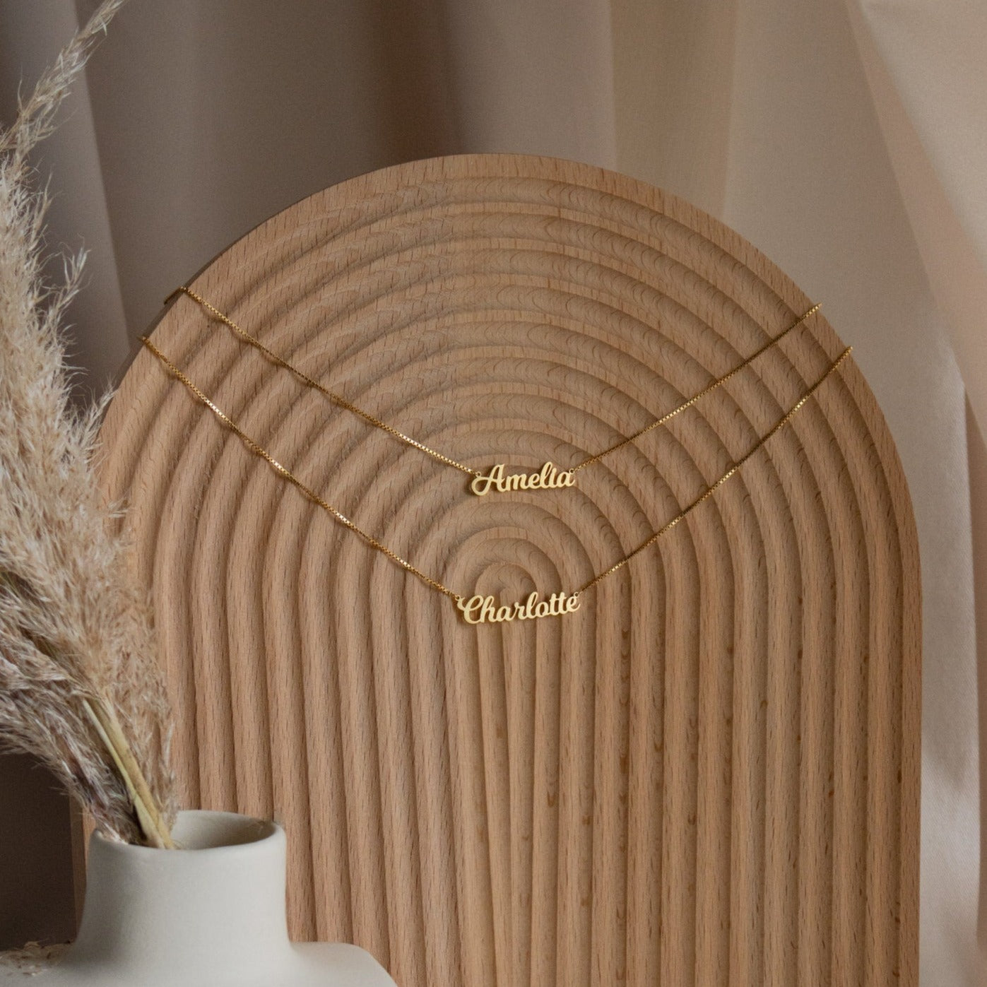 Two gold Mellow Double Name Necklaces in Box Chain, personalized with "Amelia" and "Charlotte" in a classic font, are displayed on a curved wooden stand—timeless jewelry for any occasion.
