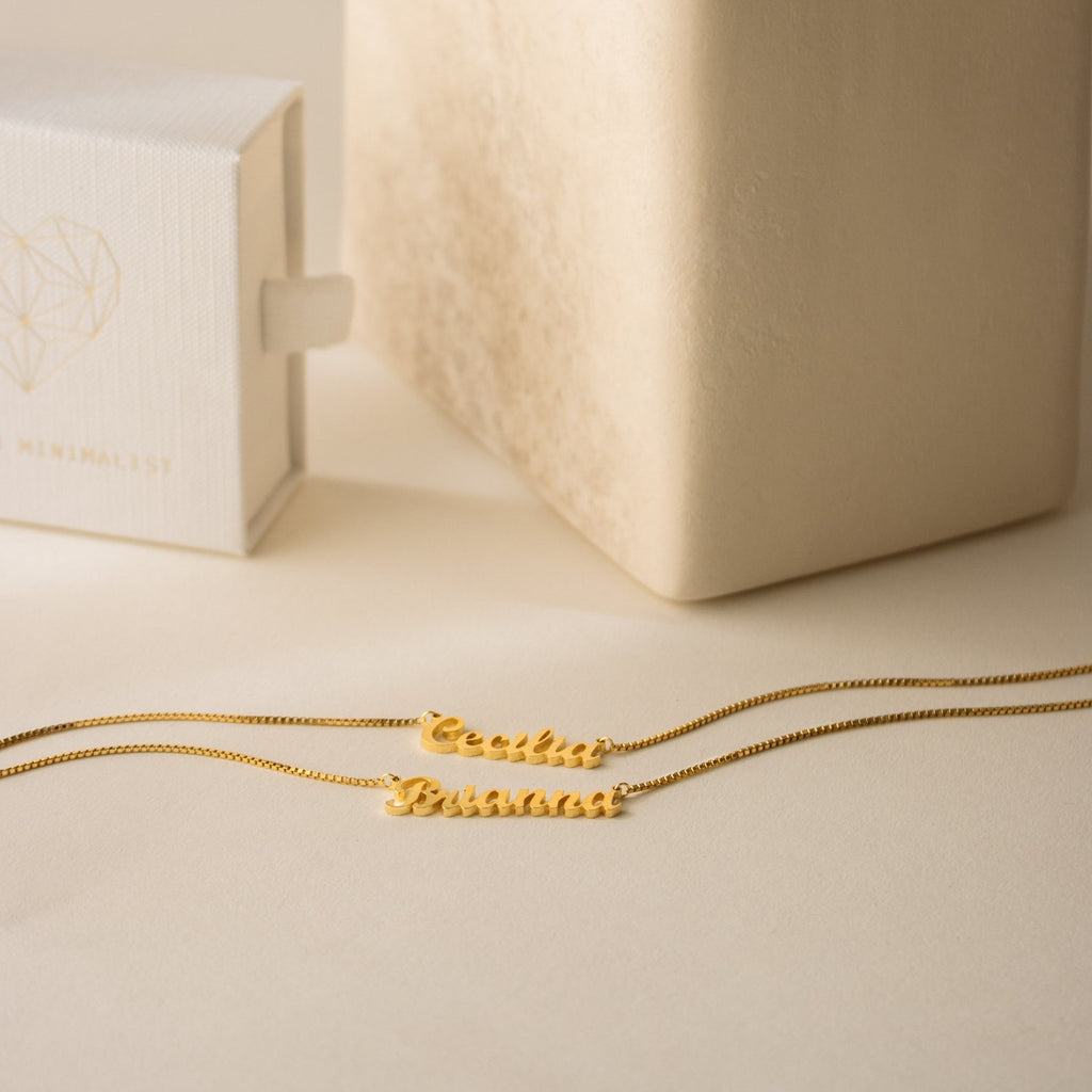 Two London Double Name Necklaces in Gold with box chains rest on a beige surface beside a white jewelry box featuring a geometric heart.