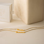 Two London Double Name Necklaces in Gold with box chains rest on a beige surface beside a white jewelry box featuring a geometric heart.