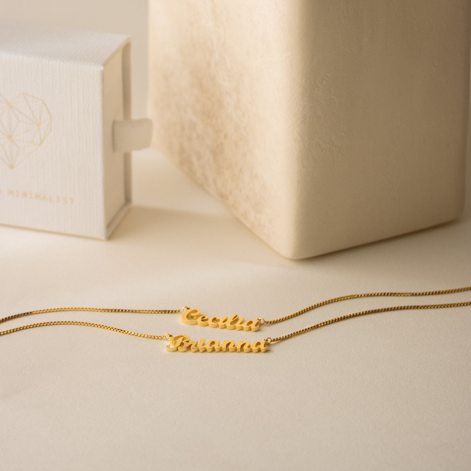 Two London Double Name Necklaces in Gold with box chains rest on a beige surface beside a white jewelry box featuring a geometric heart.