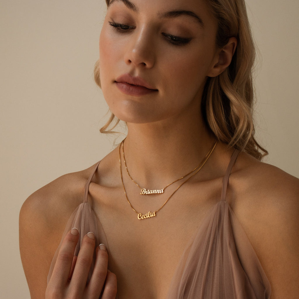 A woman wears a taupe dress and a chic London Double Name Necklace in Box Chain, personalized with "Brianna" and "Cecilia.