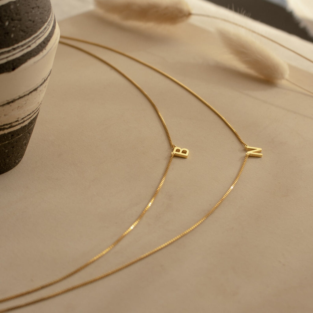 The Alice Double Letter Necklace in Box Chain features gold initial pendants "B" and "N" on a sleek box chain, displayed on a neutral surface beside a striped vase for a modern, personalized look.