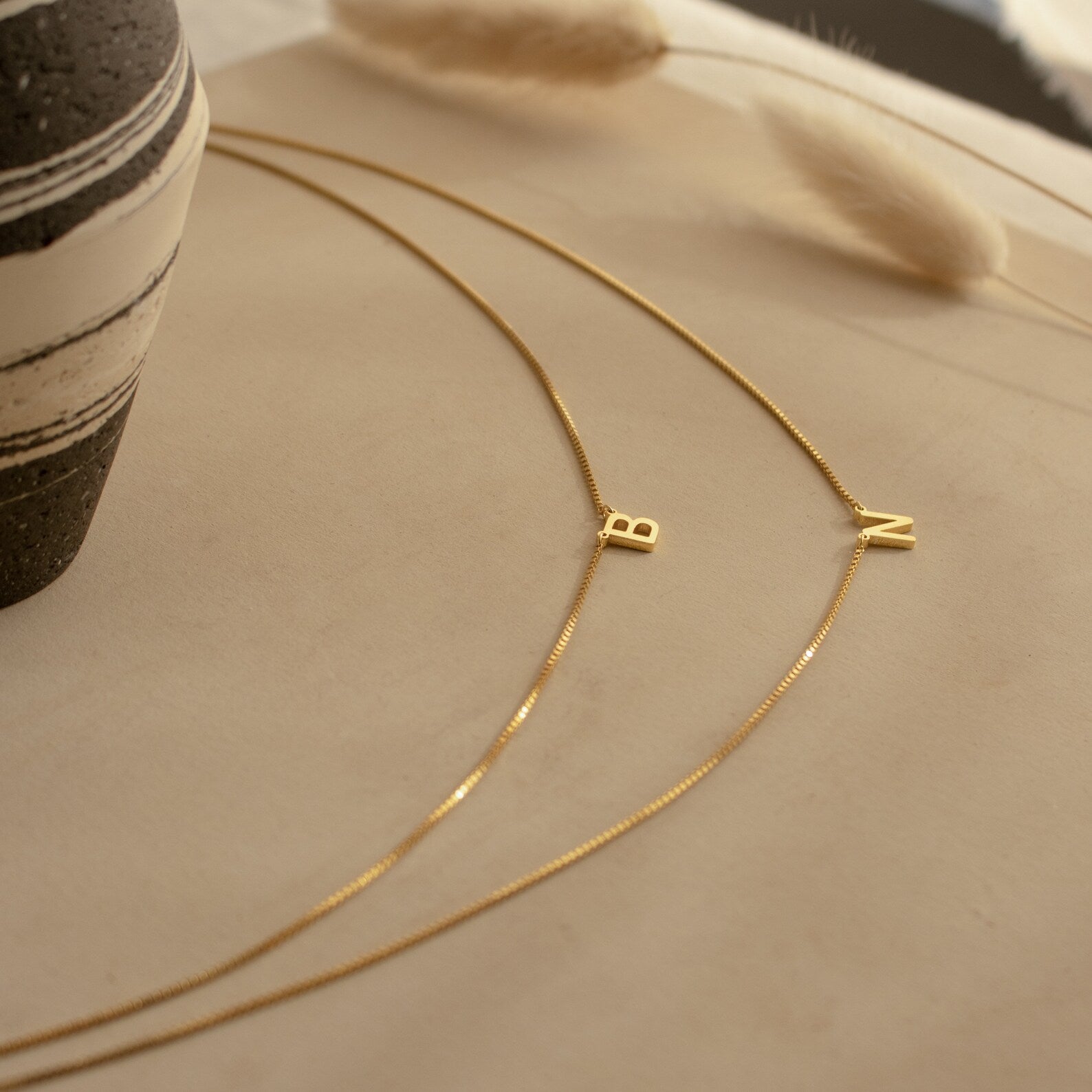 The Alice Double Letter Necklace in Box Chain features gold initial pendants "B" and "N" on a sleek box chain, displayed on a neutral surface beside a striped vase for a modern, personalized look.