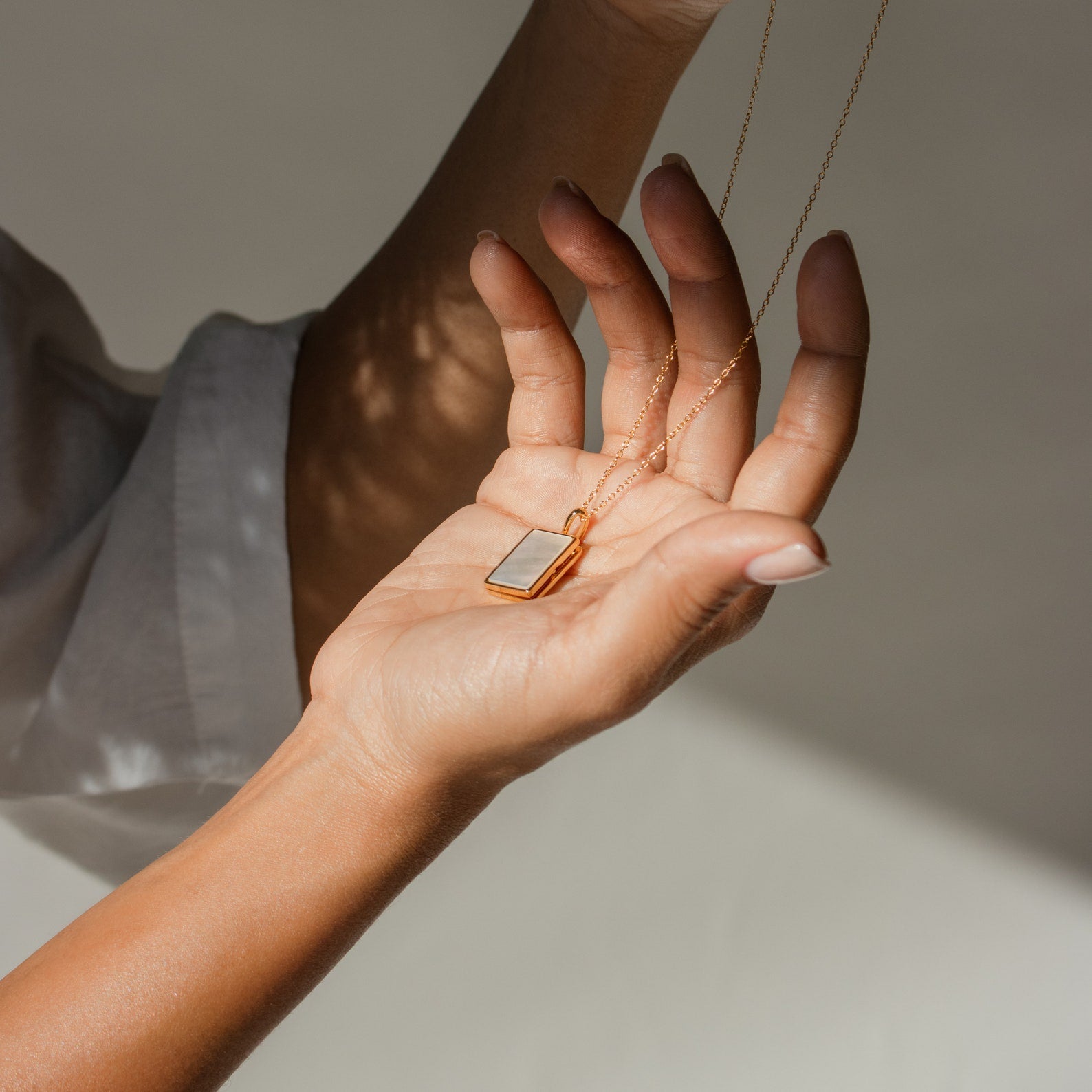 A hand holds the Pearl Locket Necklace featuring a gold, rectangular pendant, shining in natural light.
