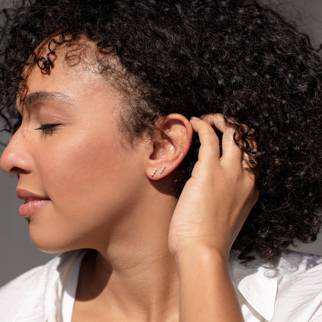 A woman with curly hair, eyes closed, touches her ear while wearing small hoops and the Tiny Pearl Bar Studs—elegant everyday earrings featuring softly glowing freshwater pearls.