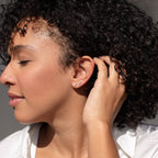 A woman with curly hair, eyes closed, touches her ear while wearing small hoops and the Tiny Pearl Bar Studs—elegant everyday earrings featuring softly glowing freshwater pearls.