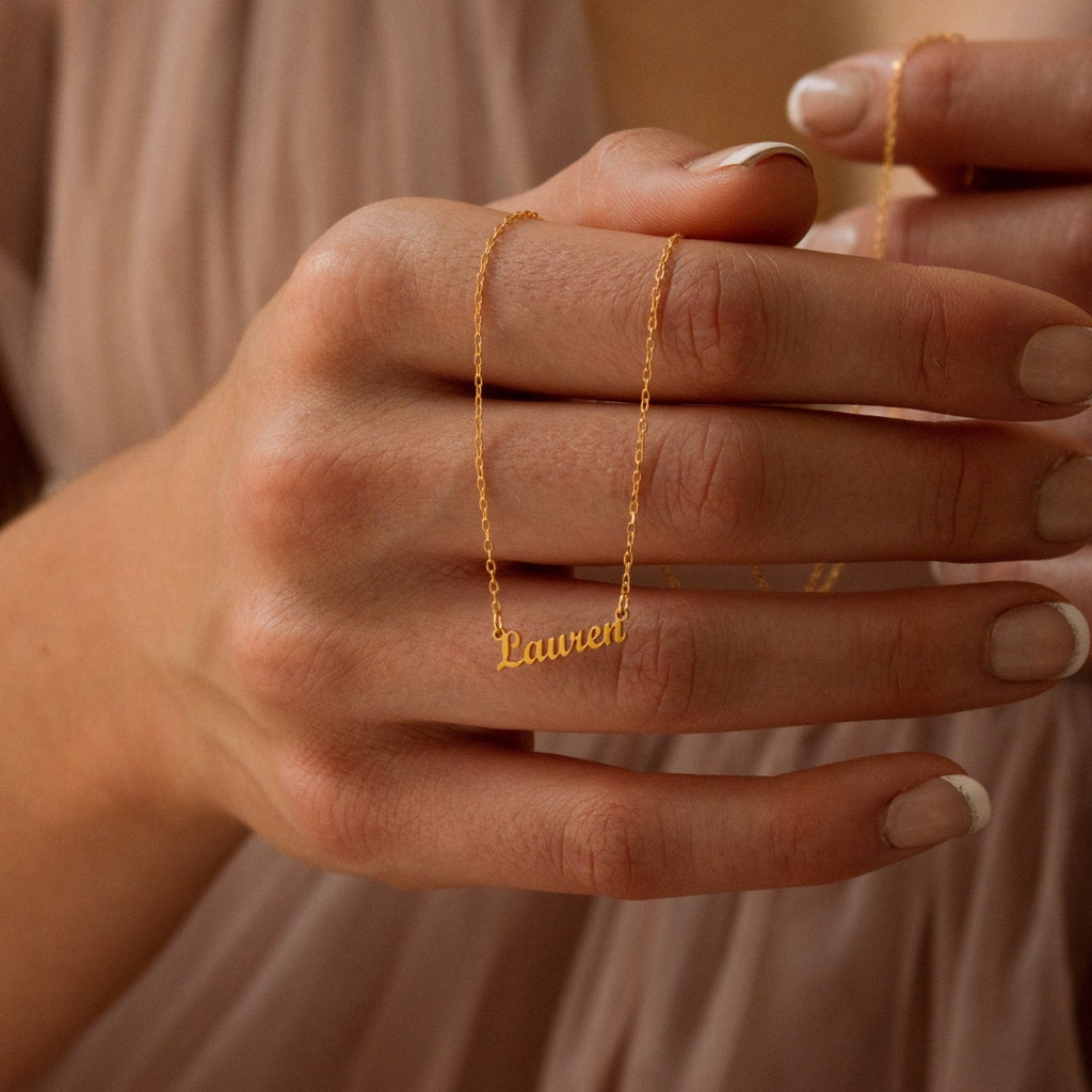 A hand displays the Tiny London Name Necklace with "Lauren" in cursive, nails done in a French manicure—ideal for fans of minimalist, customizable jewelry.