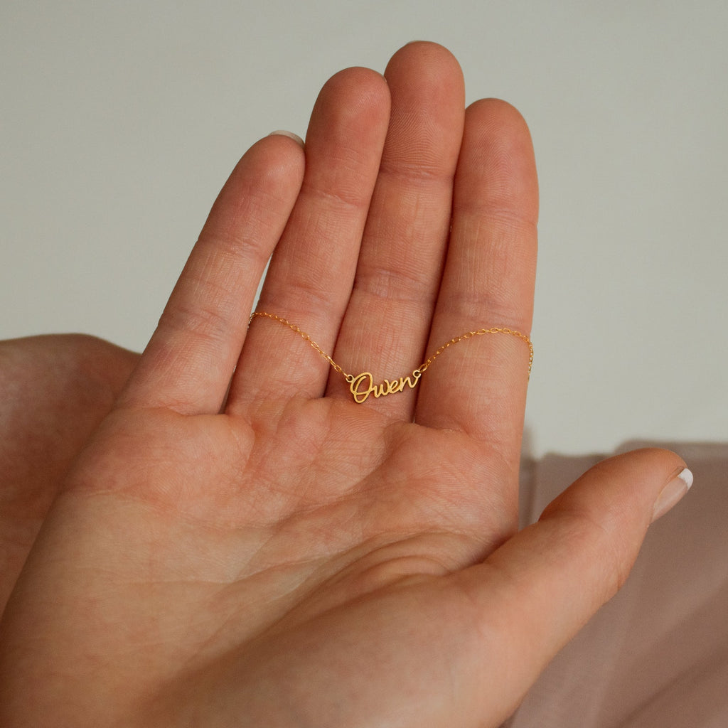 A hand holds the Tiny Grace Name Necklace, a delicate gold personalized pendant reading “Queen.”.