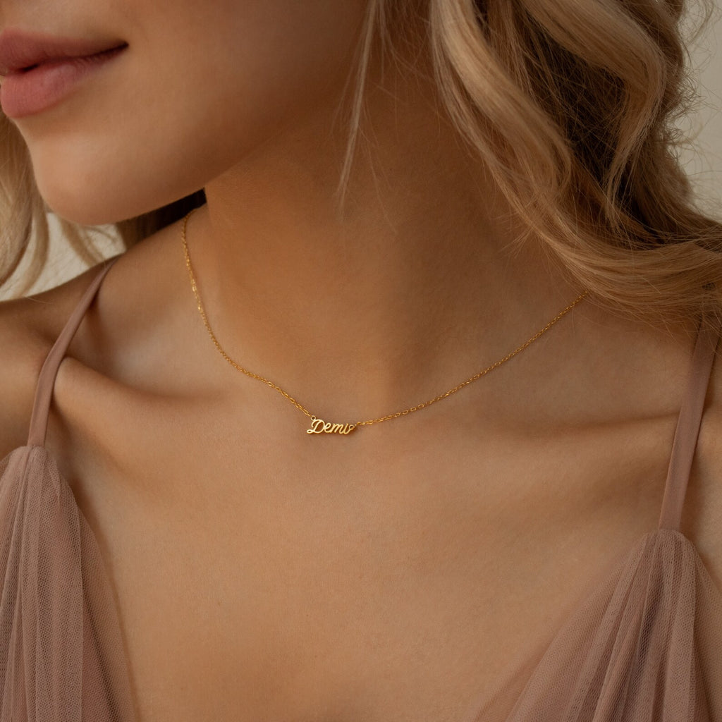 A woman wears the Tiny Paris Name Necklace in gold, featuring a Demi pendant and elegant Parisian font, paired with a sheer blush top.