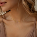 A woman wears the Tiny Paris Name Necklace in gold, featuring a Demi pendant and elegant Parisian font, paired with a sheer blush top.